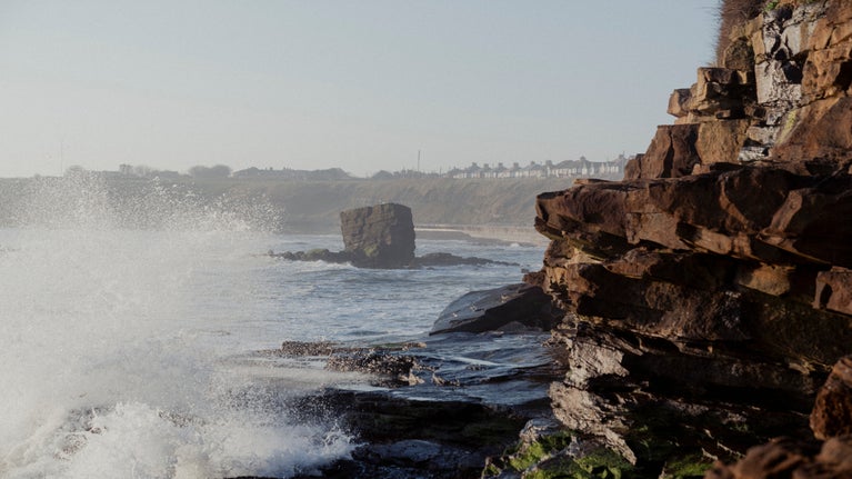 A blurred rock just off a clifftop coast line, with rocky cliff edges in focus in the foreground. There are houses on the top of the cliffs to the right, and sea spray to the left.